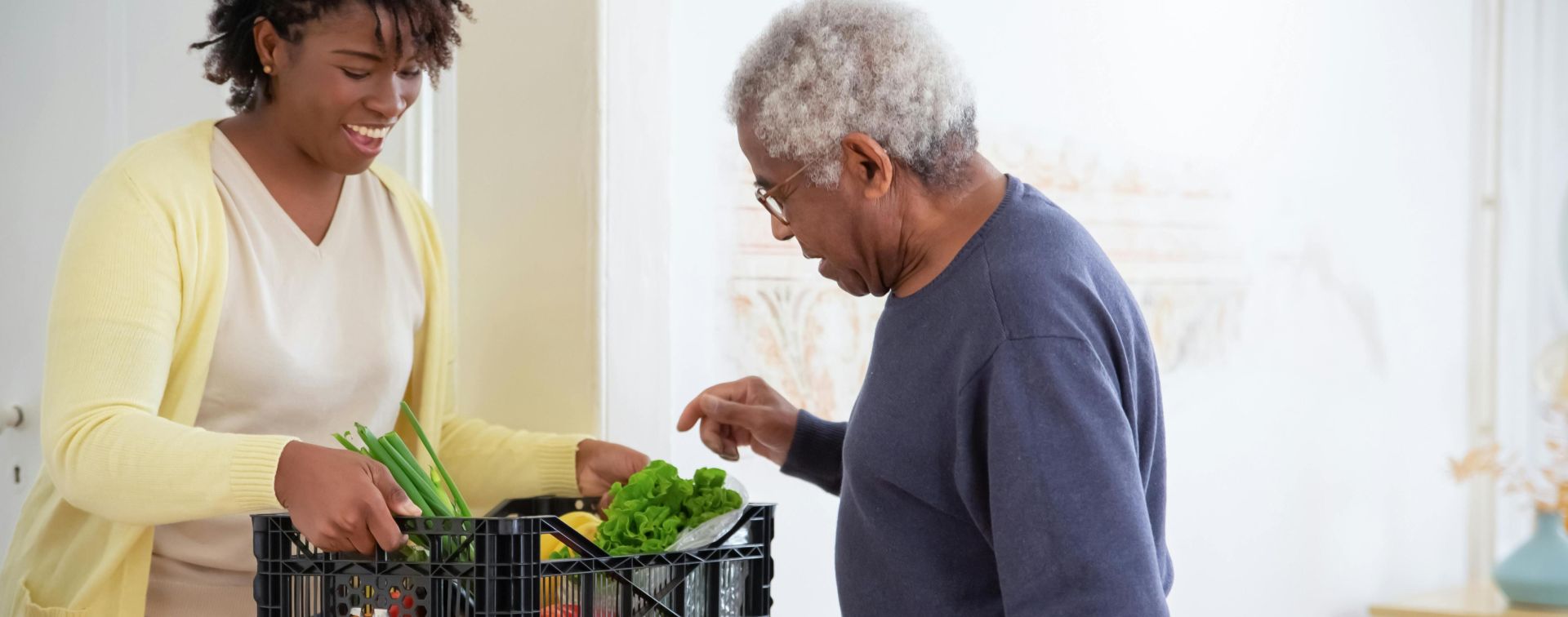 A young adult woman helps a senior man by carrying a crate of fresh vegetables indoors, symbolizing care and support.