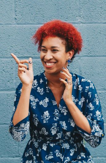 Cheerful lady with red curly hair laughing against a blue brick wall, wearing a patterned dress.